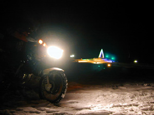 手前にバイクの輝くライトに照らされる雪原、遠く暗闇に浮かび上がる宗谷岬最北端の碑の写真