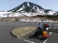 峠の駐車場で羅臼岳をバックに休んでいる荷物山盛りのバイクの写真