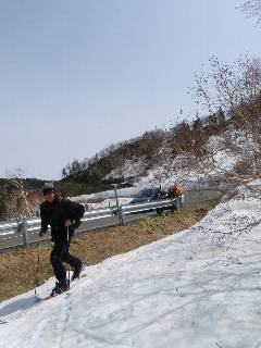道路を背景に背中にリュックサックを背負ってテレマークスキーを履いて雪の上を歩いている写真