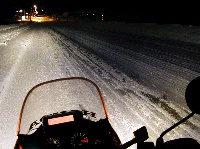 バイクのライトに照らさし出された漆黒の闇にまっすぐのびる深夜の雪道の写真