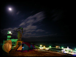 真っ暗な夜空の中に綺麗なお月様、その下に宗谷岬灯台、手前に一台のバイクとテント、遠くにライトアップされた宗谷岬の最北記念碑、全てが月に見守られている写真