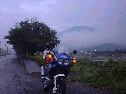 河川沿いの路、雨の中いち早く暗くなり始めた対岸の町に明かりが灯り始めている中を進むバイクの写真